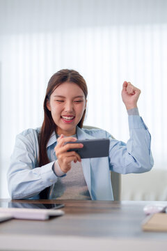 Young Asian student celebrating victory raising fist while watching smartphone at desk with copy space