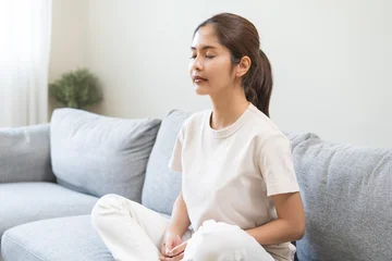 Fotobehang Lotusbloem Meditation concept, closed eye asian young woman, girl hand in calm pose sitting practice meditating in lotus position on couch at home, relax, exercise for wellbeing. Relaxation, happy leisure people  © KMPZZZ