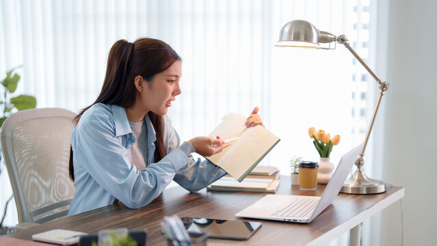 Young Asian woman reading textbook while studying with laptop at home desk with copy space