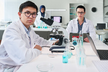 Fototapeta premium Portrait of Asian young scientist working with microscope in laboratory while female colleagues reviewing samples and tablet, supporting medical research marketing and biotech content