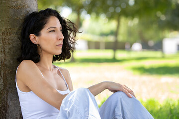 Obraz premium Thoughtful young woman sitting under a tree in a peaceful park