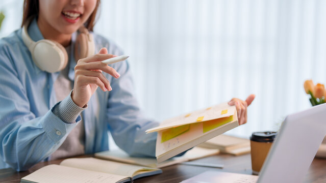 Young Asian student with headphones searching textbook pages while studying with laptop at home desk with copy space