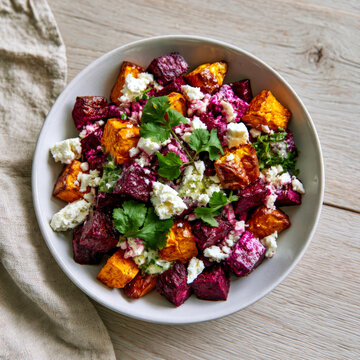 Roasted beetroot and sweet potato salad with crumbled feta cheese and fresh cilantro in white bowl on wooden table