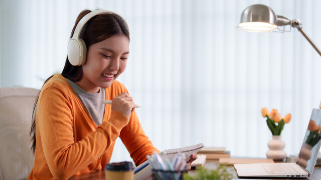 Young Asian student celebrating online learning achievement wearing headphones while studying at home desk with copy space