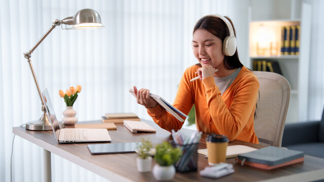 Young Asian student wearing headphones studying with notebook and computer at bright home desk for online education