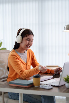 Young Asian student wearing headphones using laptop for online course at home desk with copy space