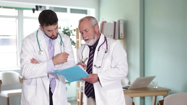 Elderly and adult male physicians stand in medical office reviewing patient test results and collaborating on diagnosis