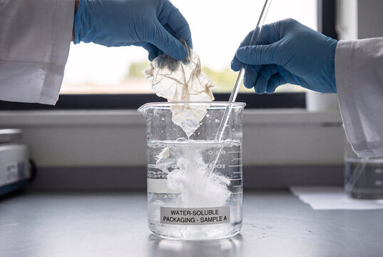 hands demonstrating a sample of water soluble packaging material dissolving quickly in a glass beaker of clear water