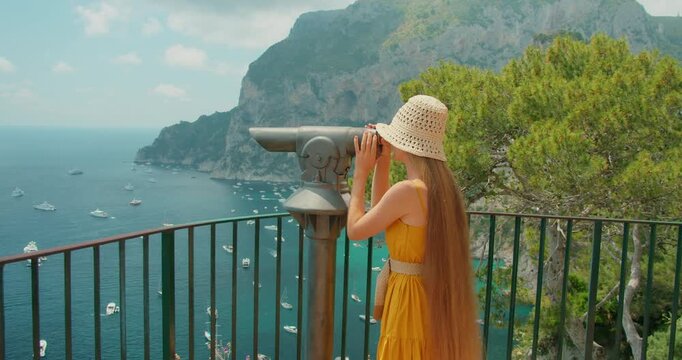 Tourist woman using viewpoint telescope over Capri cliffs and boats on calm blue sea in summer