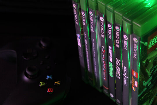 Xbox One game cases with a controller in the background, illuminated by green ambient lighting. Modern gaming setup highlighting console entertainment, collection, and technology.