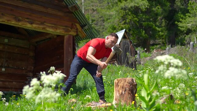 A cinematic close-up shot of an athletic male hiker in a red t-shirt splitting firewood with an axe near a wooden banya in Altai.
