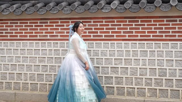 Woman in flowing blue and white hanbok stands beside traditional brick wall. Her expression is calm, eyes gently gazing ahead. Wind brushes her skirt, lifting it softly as she moves
