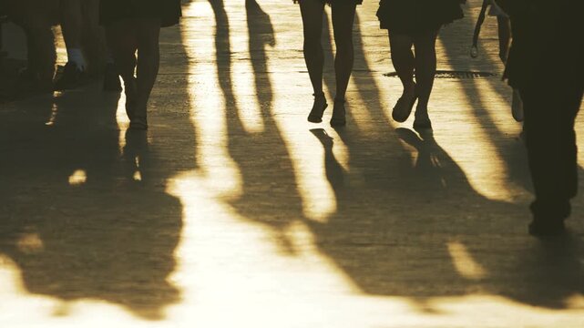 The silhouettes and shadows of people walking along the street are illuminated by the warm golden light of the setting sun, conveying the movement and atmosphere of an evening stroll.