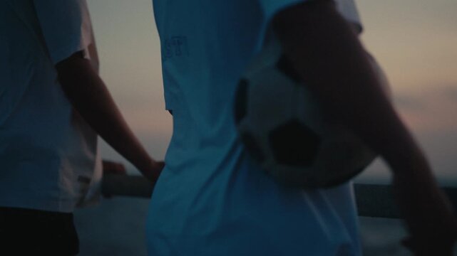 Young man holding soccer ball while standing on pier with friend near ocean at dusk