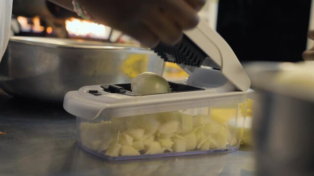 A close-up shot of a chef's hands in a professional kitchen using a manual vegetable chopper to dice fresh onions, with a fire burner visible in the background