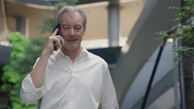Businessman phone call senior man white shirt office communication corridor professional near modern building atrium stair garden greenery smiling calm concerned expression