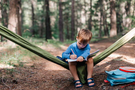 Young boy drawing in a notebook in a forest hammock
