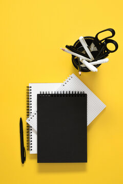 Vertical Flat Lay with Blank Black Notebook Cover, Grid Notepad, and Desk Organizer on Bright Yellow Background. Minimalist Office, Business, and Back to School Stationery Layout. Top View.