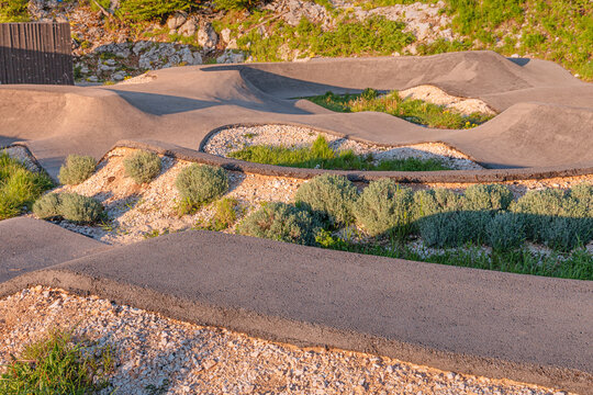 Empty asphalt pump track with berms and rollers, surrounded by gravel and green bushes, ready for biking and outdoor sports activity