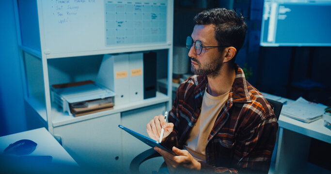 Young Caucasian male programmer use tablet for code design and plan in dark home office at night. AI technology, cybersecurity, software developer, freelance innovation, digital data network concept.