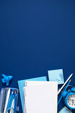 Vertical Blue Stationery Flat Lay with Blank Grid Notebook, Alarm Clock, Pencil Case, and Toy Airplane on Dark Blue Background. Back to School, Office, and Education Concept with Large Copy Space.