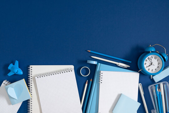 Blue Stationery Flat Lay with Notebooks, Alarm Clock, Pencils, Pen, and Toy Airplane on Dark Blue Background. Workplace, Education, and Back to School Concept Layout. Minimalist Top View Composition.