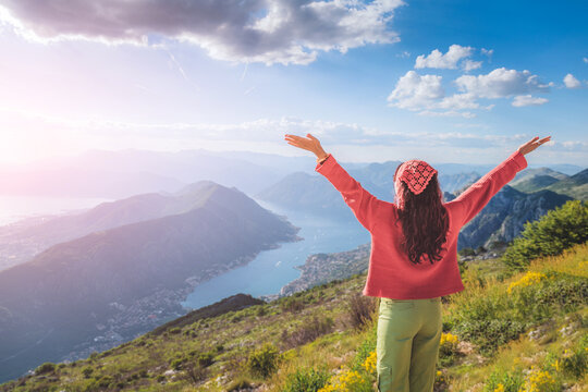 Woman standing on a mountain peak raising arms open to the sky, embracing freedom and success with a view of Bay of Kotor, Montenegro