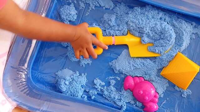 Child's hand playing with blue kinetic sand in a plastic tray using yellow shovel and pink crab mold. Sensory play activity for toddler development. Colorful toys, messy fun, and creative learning con