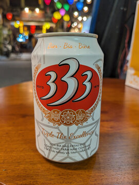 A cold can of 333 beer sits on a wooden table with colorful lights blurred in the background