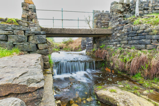 A stone bridge spans a small river with a tiered waterfall, bordered by historic stone walls and lush greenery, suggesting a scenic countryside location - Cheesden Lumb Mill, Lancashire, UK