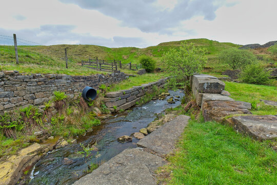 A small stream winds through green, hilly fields between ancient stone walls, with a dark pipe emerging from the left bank under a cloudy sky - Cheesden Lumb Mill, Lancashire, UK