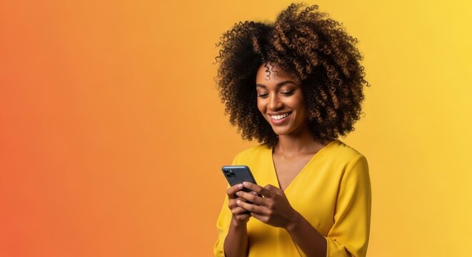 Happy young woman with curly hair looking at her smartphone against yellow background