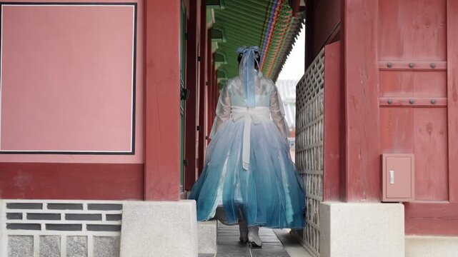 Figure in flowing blue gown walks through traditional Korean gate. Veil drapes softly as they move with quiet grace. Red wooden arches frame the ancient, serene moment. Ideal for historical, cultural