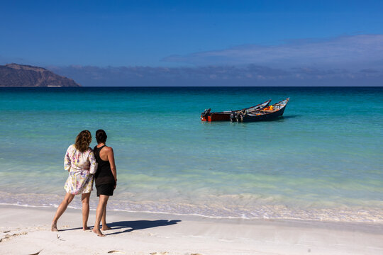 Two women standing on the white sandy beach of Shuab Beach on Socotra Island, Yemen, overlooking a traditional fishing boat in the crystal-clear turquoise sea.