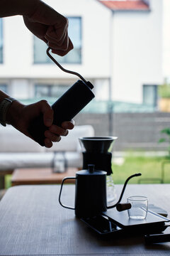 Silhouette of person grinding coffee beans with a manual grinder beside pour over coffee tools near a window. Concept of fresh coffee, hand brewing and morning routine.