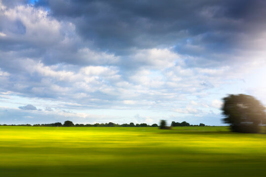 Nature in motion blur from a train window, sunlit green and yellow field with sun flare under a bright sky. Abstract travel background for design.
