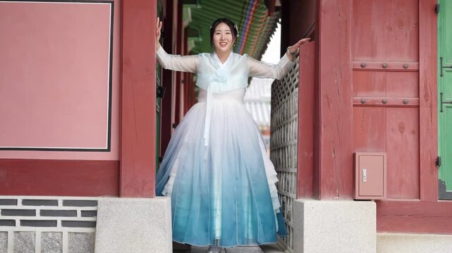 Young woman smiles, waves, steps from traditional Korean gate. She wears flowing, gradient-hued hanbok with soft white-to-blue transition