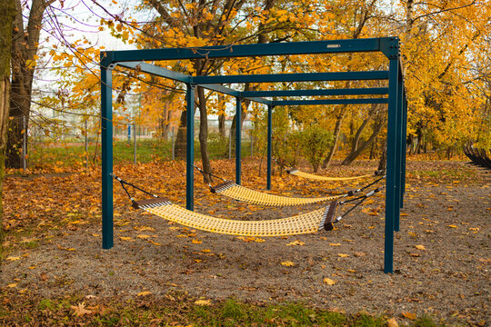 A serene outdoor hammock swing in a park setting, featuring a metal frame and fallen leaves, captured from a ground-level perspective. The scene is characterized by yellow and brown tones.
