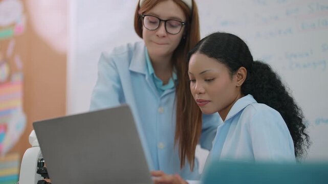 Two medical students collaborating to prepare an anatomy lesson using a laptop in a classroom equipped with a whiteboard and educational teaching materials.