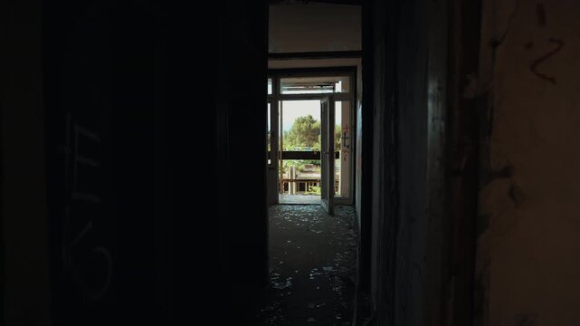 A dark corridor inside the abandoned Haludovo Hotel leads toward an open doorway with broken glass on the floor, graffiti on the walls, and daylight outside.