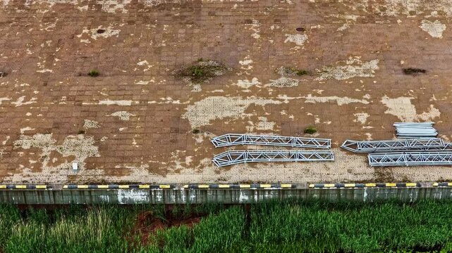 Aerial sideways shot reveals rows of metal frames and beams sorted on weathered ground beside canal at storage facility construction site