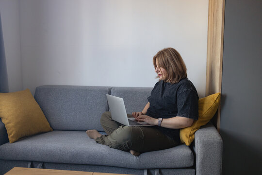 A wide shot of a young plus-size Caucasian woman sitting on a grey couch near a window, smiling at her phone screen while holding a laptop computer on her lap.