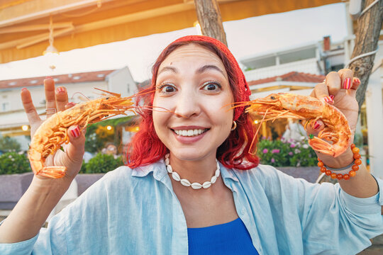 Young woman holding two large grilled shrimp, smiling at a restaurant, enjoying a funny seafood eating experience