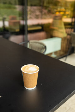 A disposable takeaway paper coffee cup with latte art stands on a wooden table with a blurred modern cafe terrace background on a summer day.