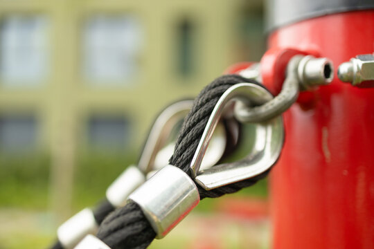 A macro close-up shot of a heavy-duty steel wire rope fastener and connector loop on a modern red metal playground climbing structure.
