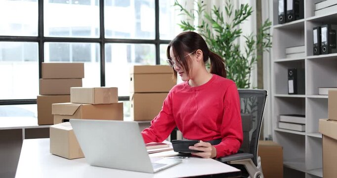 Young asian woman entrepreneur organizing customer orders at a home office, surrounded by stacks of cardboard boxes, processing online sales and managing shipping logistics for her small business