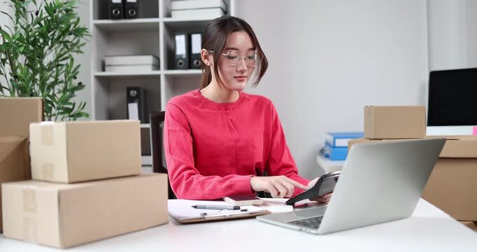 Young asian woman entrepreneur organizing customer orders at a home office, surrounded by stacks of cardboard boxes, processing online sales and managing shipping logistics for her small business