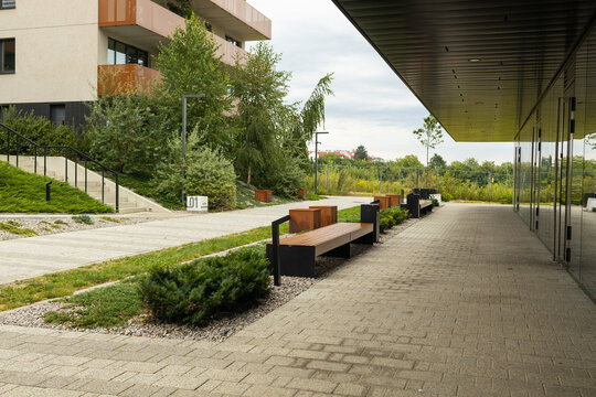 A scenic view of a contemporary residential apartment complex facade with professional courtyard landscaping, green bushes, wooden benches, and a wide brick pavement walkway.
