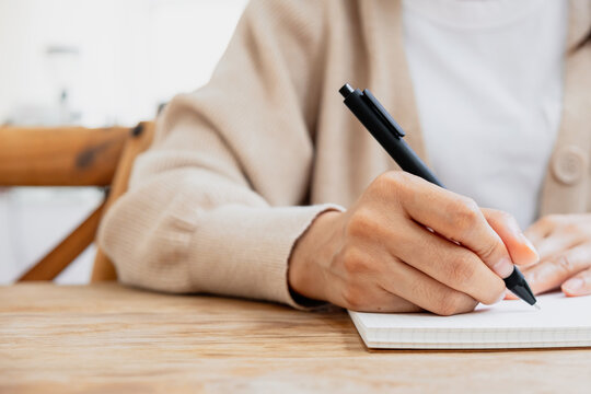 Close up of a person writing in a notebook with a pen on a wooden desk. Ideas for note taking, studying, planning and productivity concepts.