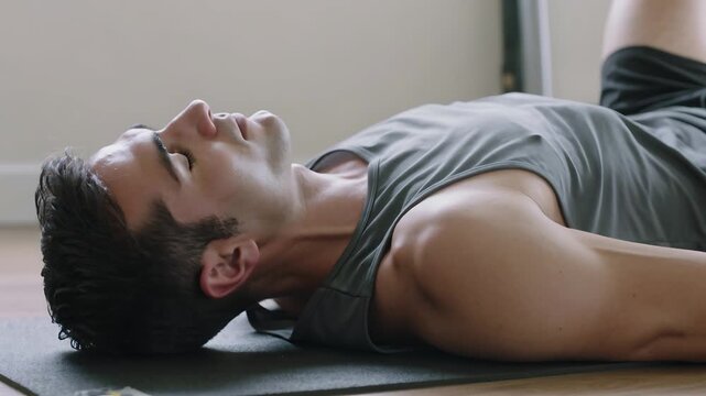 Lying man breathing slowly on dark mat on wooden floor near wall, wearing sleeveless top, shorts